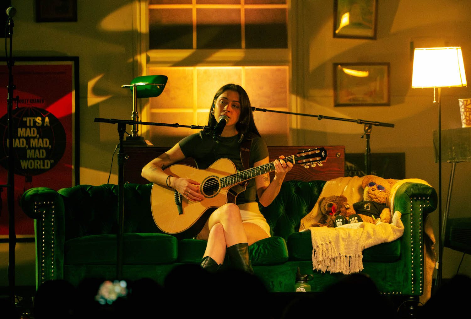 Singer seated on a green velvet couch playing an acoustic guitar on a warmly lit, living-room-style stage